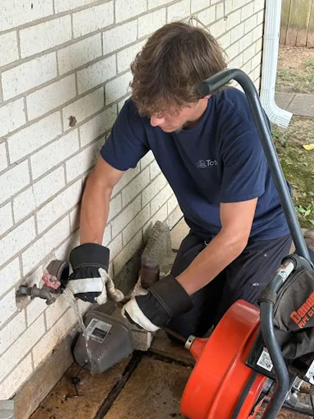 Young man using a sewer auger to clear a drain outside a brick wall.